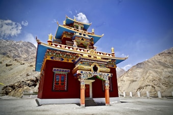 An intricately designed, colorful Buddhist monastery stands under a clear blue sky, surrounded by barren mountains. The building is adorned with vivid red, blue, gold, and green details, featuring traditional Tibetan architecture.