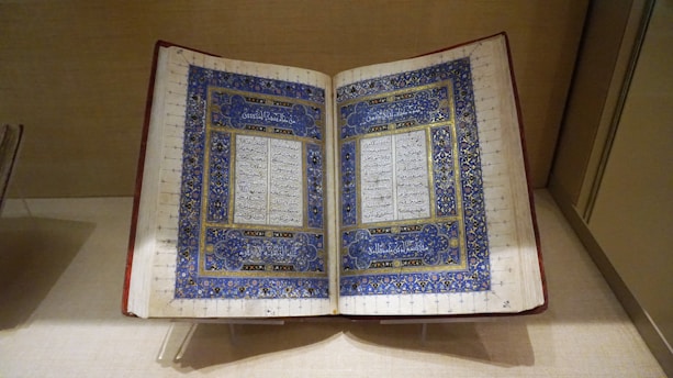 An ornate, open book is displayed on a stand, featuring intricate blue and gold decorative borders surrounding columns of handwritten text. The text appears to be in a calligraphic script, likely Arabic.
