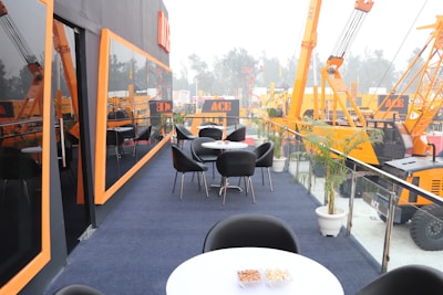 A modern outdoor seating area with round tables and black chairs is positioned on a raised platform. Large construction cranes with bright orange booms and black accents are visible in the background. Glass railings with potted plants line the edges of the seating area, which is carpeted in dark blue. The setting overlooks an industrial exhibition space, creating a blend of professional and casual atmosphere.
