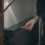 A person is inserting a key into a lock on a dark-colored panel, possibly the door of a room or piece of furniture. The person's hand is visible, wearing a watch and holding the key. Part of their arm and a suitcase handle are also visible, suggesting they are locking or unlocking something.