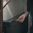 A person is inserting a key into a lock on a dark-colored panel, possibly the door of a room or piece of furniture. The person's hand is visible, wearing a watch and holding the key. Part of their arm and a suitcase handle are also visible, suggesting they are locking or unlocking something.