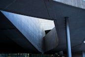 Abstract architectural structure with geometric lines and angles formed by concrete surfaces. The image captures a modern building design with visible beams and a column, emphasizing shadows and natural light.