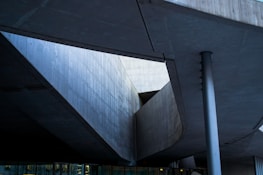 Abstract architectural structure with geometric lines and angles formed by concrete surfaces. The image captures a modern building design with visible beams and a column, emphasizing shadows and natural light.