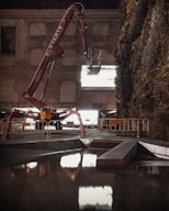 A large, red construction machine with 'ELEVATE' written on the arm is positioned in front of a brick wall. The machine is equipped with an extendable arm and reaches out towards a large window or opening that emits bright light. The surroundings include a section of reflective water in the foreground and dense foliage on the right side. Barriers line the area, suggesting a construction or renovation site.