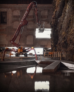 A large, red construction machine with 'ELEVATE' written on the arm is positioned in front of a brick wall. The machine is equipped with an extendable arm and reaches out towards a large window or opening that emits bright light. The surroundings include a section of reflective water in the foreground and dense foliage on the right side. Barriers line the area, suggesting a construction or renovation site.