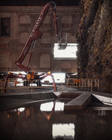 A large, red construction machine with 'ELEVATE' written on the arm is positioned in front of a brick wall. The machine is equipped with an extendable arm and reaches out towards a large window or opening that emits bright light. The surroundings include a section of reflective water in the foreground and dense foliage on the right side. Barriers line the area, suggesting a construction or renovation site.