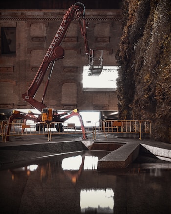 A large, red construction machine with 'ELEVATE' written on the arm is positioned in front of a brick wall. The machine is equipped with an extendable arm and reaches out towards a large window or opening that emits bright light. The surroundings include a section of reflective water in the foreground and dense foliage on the right side. Barriers line the area, suggesting a construction or renovation site.