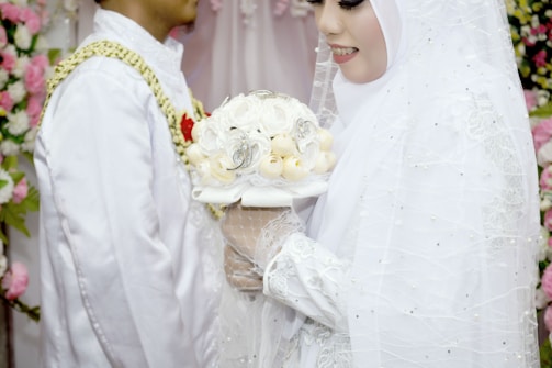 A bride wearing a white hijab and a detailed white dress holds a bouquet of white flowers adorned with intricate decorations. A groom stands next to the bride, wearing a white traditional outfit with gold and red accents. The background is decorated with an array of pink and white flowers.