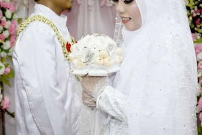 A bride wearing a white hijab and a detailed white dress holds a bouquet of white flowers adorned with intricate decorations. A groom stands next to the bride, wearing a white traditional outfit with gold and red accents. The background is decorated with an array of pink and white flowers.