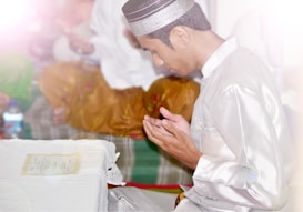 A person dressed in traditional attire sits with hands raised in a gesture of prayer or meditation. Nearby, there is a table covered with a white cloth and a piece of paper placed on it. The lighting is soft, with a warm, serene atmosphere.