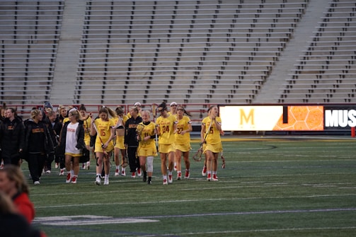 A group of athletes in yellow uniforms, holding lacrosse sticks, walks across a sports field. They are accompanied by coaches or team members in black jackets. The stands in the background are empty, and a digital scoreboard displays part of a letter and some graphics.