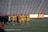 A group of athletes in yellow uniforms, holding lacrosse sticks, walks across a sports field. They are accompanied by coaches or team members in black jackets. The stands in the background are empty, and a digital scoreboard displays part of a letter and some graphics.