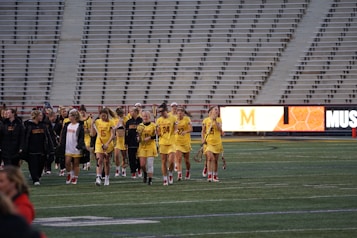 A group of athletes in yellow uniforms, holding lacrosse sticks, walks across a sports field. They are accompanied by coaches or team members in black jackets. The stands in the background are empty, and a digital scoreboard displays part of a letter and some graphics.