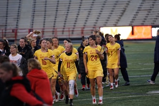 A group of female athletes wearing yellow uniforms with red and black accents walk together on a sports field. They appear to be part of a team, possibly celebrating or regrouping after a game. A large empty stadium with seating is visible in the background.