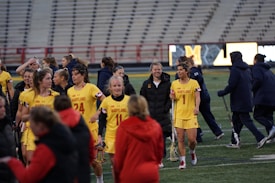 A group of female athletes in yellow uniforms, featuring the word 'Maryland' and numbers, are gathered on a sports field. Some are holding lacrosse sticks, and they appear to be moving as a group, possibly after a game. Surrounding them are a few people in dark jackets. The background shows stadium seating and an electronic display board.