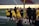 A group of athletes wearing yellow sports uniforms are standing on a sports field. Some are holding lacrosse sticks, and they appear to be interacting casually. Several people are dressed in black coats. In the background, there is a bright sign with text and parts of a stadium are visible.