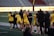 A group of athletes wearing yellow sports uniforms are standing on a sports field. Some are holding lacrosse sticks, and they appear to be interacting casually. Several people are dressed in black coats. In the background, there is a bright sign with text and parts of a stadium are visible.