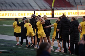 A group of athletes wearing yellow sports uniforms are standing on a sports field. Some are holding lacrosse sticks, and they appear to be interacting casually. Several people are dressed in black coats. In the background, there is a bright sign with text and parts of a stadium are visible.