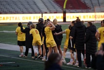 A group of athletes wearing yellow sports uniforms are standing on a sports field. Some are holding lacrosse sticks, and they appear to be interacting casually. Several people are dressed in black coats. In the background, there is a bright sign with text and parts of a stadium are visible.