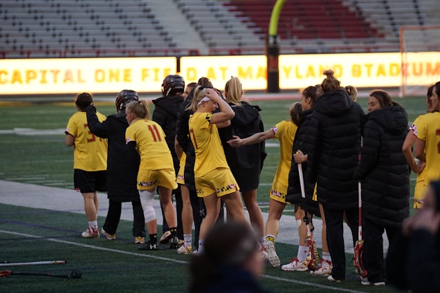 A group of athletes wearing yellow sports uniforms are standing on a sports field. Some are holding lacrosse sticks, and they appear to be interacting casually. Several people are dressed in black coats. In the background, there is a bright sign with text and parts of a stadium are visible.