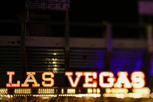 Bold, illuminated letters spell out 'Las Vegas' against a dark background, with vibrant colors and a blurred backdrop suggesting a lively, bustling environment.