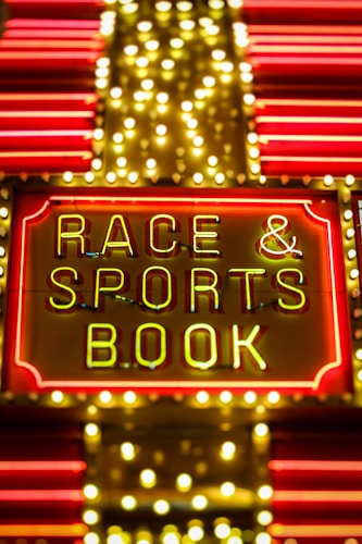 Bright neon lights on a marquee sign display the words 'RACE & SPORTS BOOK'. The sign is surrounded by rows of yellow and white lights against a red background, evoking a vibrant, energetic atmosphere typically associated with casinos or entertainment venues.