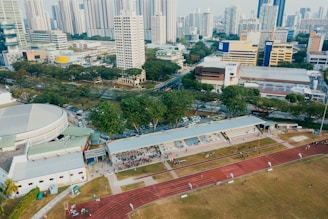 An aerial view of an urban area featuring a sports complex with a track field and a grandstand. Surrounding the sports area are numerous high-rise buildings, greenery, and roads with vehicles. The image captures people engaging in activities on the field.