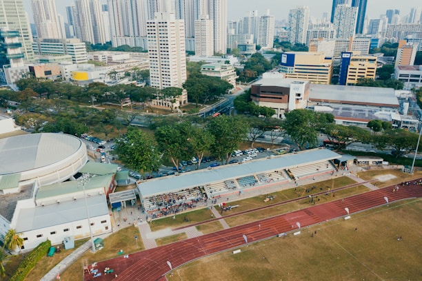 An aerial view of an urban area featuring a sports complex with a track field and a grandstand. Surrounding the sports area are numerous high-rise buildings, greenery, and roads with vehicles. The image captures people engaging in activities on the field.