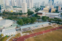 An aerial view of an urban area featuring a sports complex with a track field and a grandstand. Surrounding the sports area are numerous high-rise buildings, greenery, and roads with vehicles. The image captures people engaging in activities on the field.