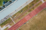An aerial view of a sports complex featuring a running track and a grassy field. The tracks are red and situated next to a large building. There are several people scattered across the area, some on the track and others near the building. The surroundings include trees and geometric pathways, adding structure to the scene.