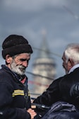 Two elderly men are engaged in what appears to be a conversation or shared activity. The man in the foreground wears a brown hat and dark jacket with a yellow stripe. He has a thoughtful expression, while the other man, seen from behind, has white hair and a dark jacket. The background shows a blurred structure, possibly a tower or building.