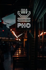 A dimly lit street scene featuring a neon sign with the word 'PHO' and a bowl design above it. The sign is mounted on a building exterior, while below, there are wet tables and benches, indicating recent rainfall. The background shows pedestrians walking along the street, with additional warm lighting from street lamps.