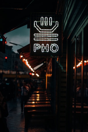 A dimly lit street scene featuring a neon sign with the word 'PHO' and a bowl design above it. The sign is mounted on a building exterior, while below, there are wet tables and benches, indicating recent rainfall. The background shows pedestrians walking along the street, with additional warm lighting from street lamps.