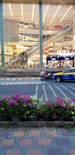 A bustling urban scene featuring a modern multi-story shopping mall with large glass windows. Shoppers can be seen on escalators and through the windows, indicating a lively interior. In front of the mall, there's a road with a blue and yellow taxi and a motorcycle carrying two people. The foreground displays colorful pink, purple, and white flowers in pots lining the sidewalk.