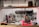 A professional kitchen setting with organized shelves of spices and condiments, various cooking utensils hanging on the wall, and a commercial oven. Copper pendant lights hang from the ceiling, and there are stacks of plates and bowls on the counter. Coffee mugs are neatly arranged on one side.