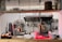 A professional kitchen setting with organized shelves of spices and condiments, various cooking utensils hanging on the wall, and a commercial oven. Copper pendant lights hang from the ceiling, and there are stacks of plates and bowls on the counter. Coffee mugs are neatly arranged on one side.