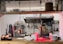 A professional kitchen setting with organized shelves of spices and condiments, various cooking utensils hanging on the wall, and a commercial oven. Copper pendant lights hang from the ceiling, and there are stacks of plates and bowls on the counter. Coffee mugs are neatly arranged on one side.