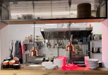 A professional kitchen setting with organized shelves of spices and condiments, various cooking utensils hanging on the wall, and a commercial oven. Copper pendant lights hang from the ceiling, and there are stacks of plates and bowls on the counter. Coffee mugs are neatly arranged on one side.