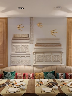 A dining area with a long wooden table set with dishes containing assorted foods. The table is arranged with plates, cups, and napkins. There are decorative pillows on a beige cushioned seating, displaying a variety of colors and patterns. The wall features ornate designs and three golden bird sculptures, creating an elegant visual display.