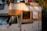 A vintage camper van parked in a natural setting with trees casting shadows on its exterior. The vehicle's side is painted in white and orange, and it has clear signs of wear and aging. The roof is loaded with items including a red bag and a plastic crate.