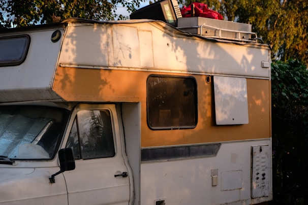 A vintage camper van parked in a natural setting with trees casting shadows on its exterior. The vehicle's side is painted in white and orange, and it has clear signs of wear and aging. The roof is loaded with items including a red bag and a plastic crate.