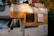 A vintage camper van parked in a natural setting with trees casting shadows on its exterior. The vehicle's side is painted in white and orange, and it has clear signs of wear and aging. The roof is loaded with items including a red bag and a plastic crate.