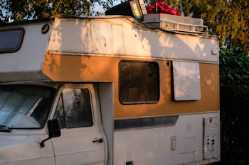A vintage camper van parked in a natural setting with trees casting shadows on its exterior. The vehicle's side is painted in white and orange, and it has clear signs of wear and aging. The roof is loaded with items including a red bag and a plastic crate.