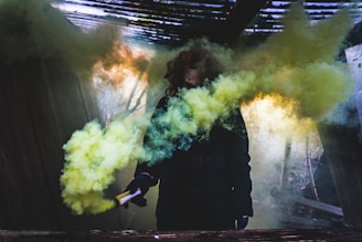 A person holds a smoke bomb releasing thick yellow and green smoke in a dimly lit environment. The surroundings appear to be an abandoned structure with wooden elements and a partially open ceiling allowing light to filter through, casting eerie shadows.