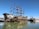 A large, intricately designed pirate ship features multiple sails and a decorative stern with a pirate motif. It is docked on calm waters alongside other smaller ships under a clear blue sky.