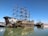 A large, intricately designed pirate ship features multiple sails and a decorative stern with a pirate motif. It is docked on calm waters alongside other smaller ships under a clear blue sky.
