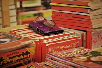 A collection of books stacked on a shelf, primarily featuring vibrant red and orange covers. Among the books, a pair of reading glasses and a purple case are placed on top of the stack.