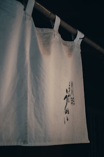 A white curtain-like fabric with Japanese characters printed in black ink hangs from a wooden rod. The fabric is slightly wrinkled and appears to have a traditional, minimalist design.