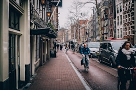 A busy urban street with people walking and cycling alongside parked cars. The architecture is characterized by European-style buildings with narrow facades and large windows. The atmosphere is lively with a mix of pedestrians and cyclists navigating the street.
