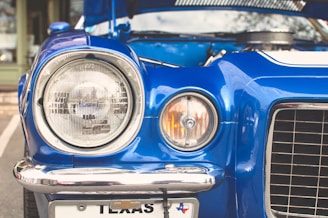 A close-up view of the front of a classic blue car, featuring round headlights and an open hood. Chrome accents enhance the vintage feel, with a Texas license plate visible at the bottom.